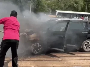 कार से उठती आग की लपटें और पाइप से आग बुझाने का प्रयास करते व्यक्ति।