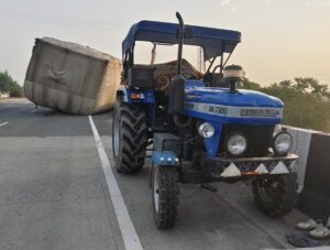 बिना नंबर प्लेट और ओवरलोड ट्रैक्टर-ट्रॉली हादसे के बाद हाईवे किनारे फंसी, सड़क सुरक्षा पर उठे सवाल।